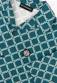 Teal kortemouwen shirt met een wit geometrisch patroon, zwarte logo patch en knopen. Materiaal lijkt zacht en licht textuur te hebben.