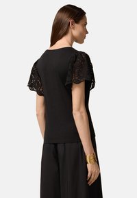 Black t-shirt with embroidered lace sleeves, round neckline, and a fitted design. Paired with loose black trousers and a gold cuff bracelet.