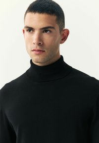 Crni turtleneck pulover od glatkog, rastezljivog materijala s uklasnim dizajnom i rebrastim ovratnikom. Prikaz izbliza ovratnika i ramena.