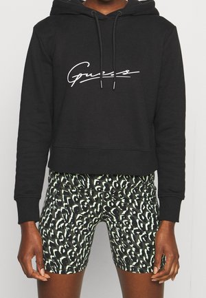 Personne portant un sweat à capuche court noir avec une écriture blanche et un short à motif abstrait noir et blanc, debout devant un fond uni.