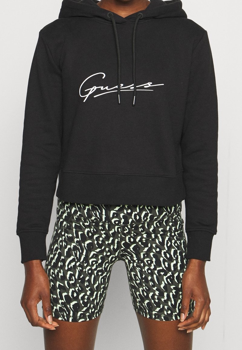 Personne portant un sweat à capuche court noir avec une écriture blanche et un short à motif abstrait noir et blanc, debout devant un fond uni.