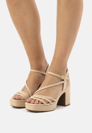 Tacones de plataforma de ante beige con tiras entrelazadas, punta cuadrada y tacón grueso. Cierre de hebilla en la correa del tobillo. Textura suave.