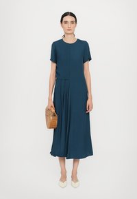 Syvän teal-värinen midi-mekko lyhyillä hihoilla, laskostettu hame, pyöreä pääntie, yhdistettynä kudottuun beigeen käsilaukkuun ja kermanvärisiin kärjellisiin kenkiin.