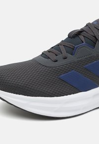 Grey athletic sneaker with a textured mesh upper, navy accents, and a white cushioned sole. Features a lace-up closure and reinforced toe area.