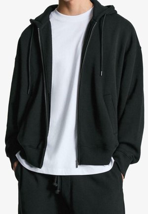 Hombre con sudadera negra con cremallera sobre camiseta blanca, con las manos en los bolsillos de pantalones deportivos negros, recortado en el cuello y las caderas.