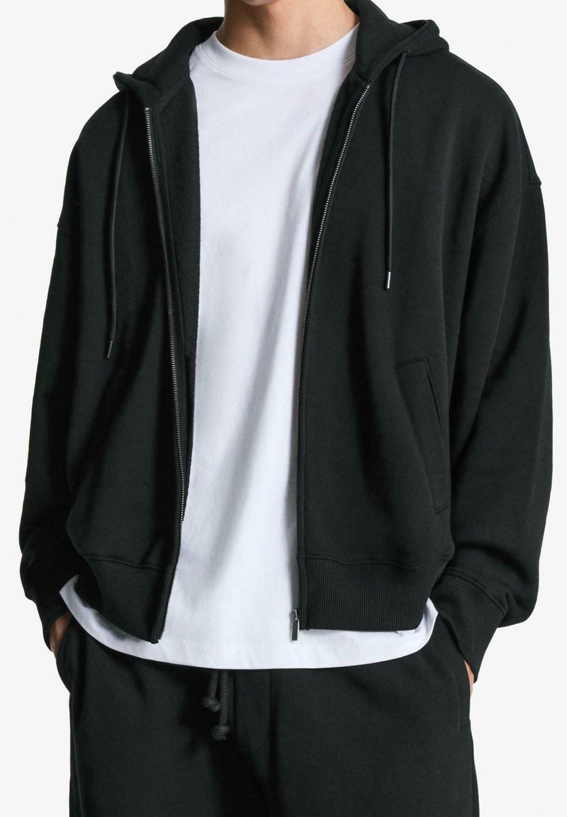 Homme portant un sweat à capuche noir zippé par-dessus un t-shirt blanc, les mains dans les poches d'un pantalon de jogging noir, cadré au niveau du cou et des hanches.