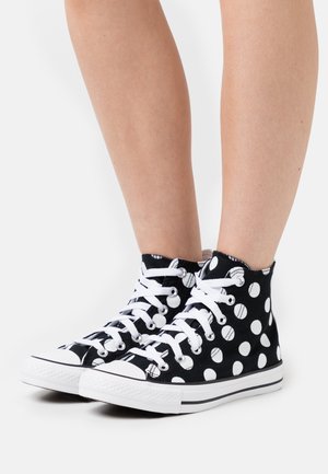 Černé vysoké tenisky s velkými bílými puntíky, bílými šňůrkami a bílou gumovou podrážkou, nošené na bosých nohou.