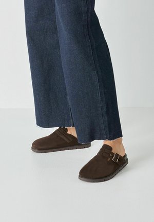 Chaussures marron en daim à enfiler avec deux brides à boucle, associées à un pantalon en jean bleu à jambes larges avec ourlets effilochés.