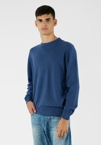 Niebieski sweter dzianinowy z okrągłym dekoltem, ściągaczami przy mankietach i na dole. Noszony z jasnoniebieskimi dżinsami. Gładka faktura, minimalistyczny design i brak widocznych wzorów.