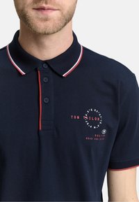 Marineblå polo med en rød og hvidstribet krave, tre-knap placket og fronttryk med teksten "Tom Tailor" samt nautiske designelementer.