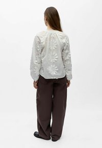 Femme debout de dos portant un chemisier blanc brodé de fleurs et un pantalon ample marron foncé avec des chaussures noires.