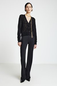 Schwarzer Strickcardigan mit Rautenmuster, Knopfleiste, kombiniert mit maßgeschneiderten schwarzen Hosen und spitzen schwarzen Schuhen, ergänzt durch eine auffällige goldene Kette.