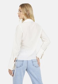 Camicia bianca a maniche lunghe con colletto, con dettagli in cravatta sulle maniche e tessuto testurizzato. Abbinata a jeans a vita alta di colore blu chiaro.