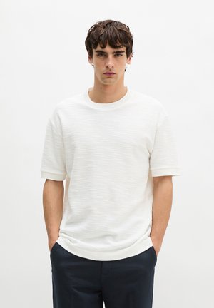 Junger Mann mit dunklem, lockigem Haar, der einen strukturierten weißen Kurzarmpullover und dunkle Hosen trägt, mit den Händen in den Taschen vor einem weißen Hintergrund stehend.