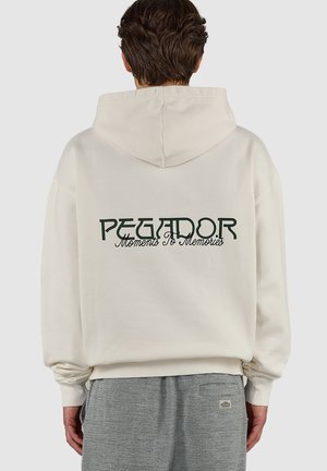 Osoba ubrana w kremowy kaptur z napisem „PEGADOR Moments to Memories” na plecach oraz szare spodnie w jodełkę, zwrócona tyłem.