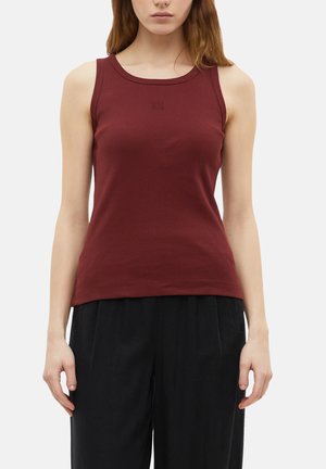 Femme portant un débardeur côtelé bordeaux sans manches et un pantalon noir ample, debout devant un fond clair uni.