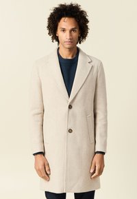 Abrigo de mezcla de lana color beige con solapas muescas, dos botones y bolsillos laterales. Llevado sobre un suéter azul oscuro, que presenta una silueta ajustada.
