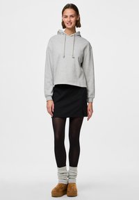 Jeune femme debout, portant un sweat à capuche court gris, une mini-jupe noire, des collants noirs, des guêtres grises et des chaussures à plateforme marron.