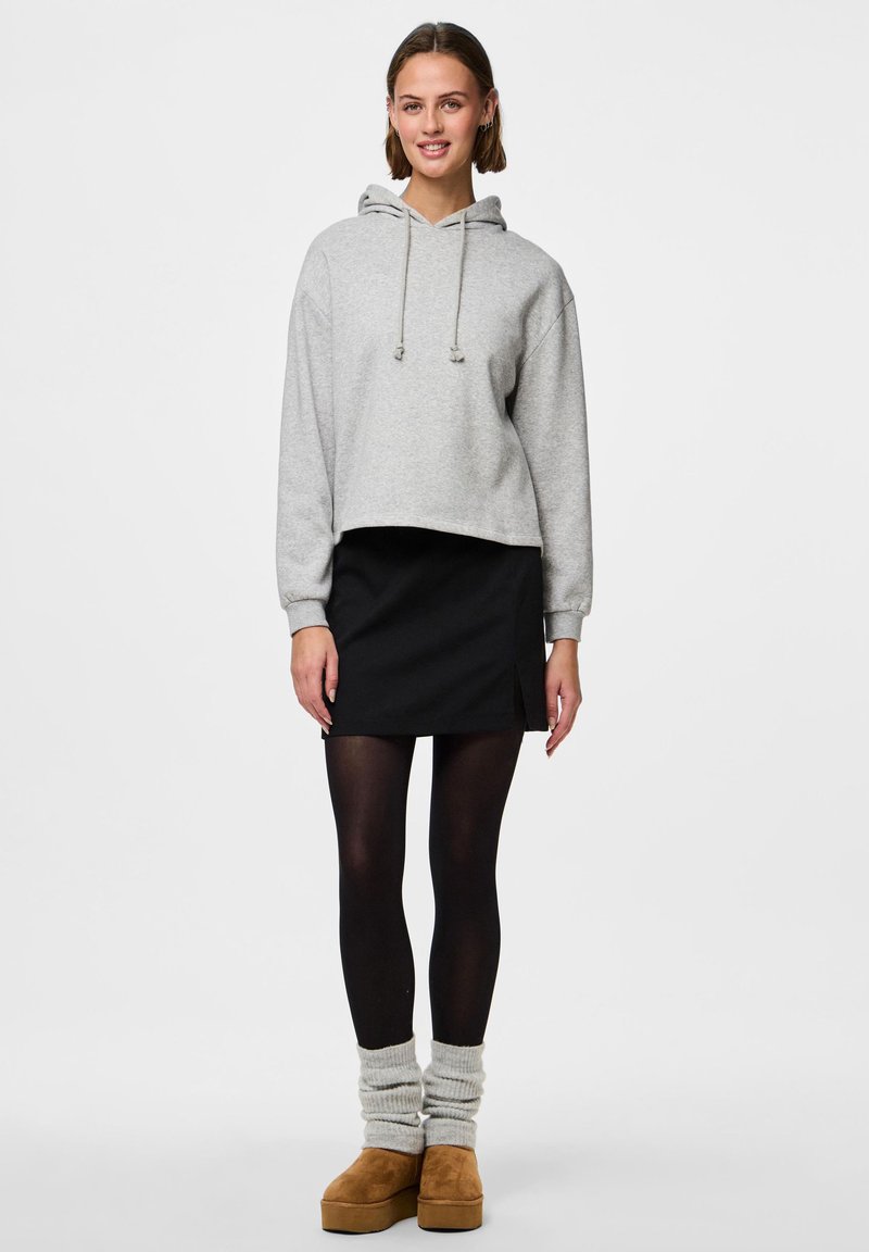 Jeune femme debout, portant un sweat à capuche court gris, une mini-jupe noire, des collants noirs, des guêtres grises et des chaussures à plateforme marron.