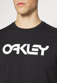 Sort bomulds t-shirt med et stort, fedt hvidt "OAKLEY" logo på brystet. Enkel design med rund hals og en glat tekstur.