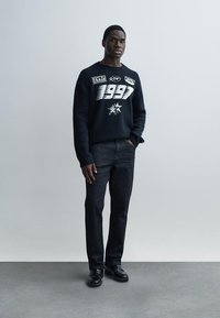 Schwarzer Pullover mit Grafikdruck und weißen Akzenten, kombiniert mit dunkler Jeans. Modell steht vor einem schlichten Hintergrund.