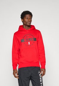 Ung mann med rød Chicago Bulls hettegenser og svarte Bulls sportbukser, stående mot en ensfarget lys bakgrunn.