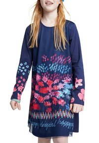 Enfant portant une robe à manches longues bleu marine avec des motifs floraux colorés en rose, rouge et bleu, debout devant un fond blanc.