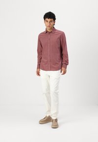 Langärmliges, knöpfbares Hemd aus staubiger Rose, kombiniert mit weißen Slim-Fit-Hosen. Das Model steht vor einem neutralen Hintergrund und trägt beiger Schuhe.