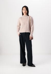 Világos rózsaszín kötött pulóver magas nyakkivágással és lyukacsos mintával, sötét, bő szárú nadrággal és fekete hegyes orrú cipővel párosítva.