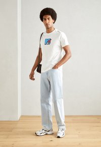 Camiseta blanca de algodón con el logo de MTV en azul y rojo, jeans rectos de mezclilla azul claro y zapatillas plateadas con detalles en negro. Bolso marrón bandolera.