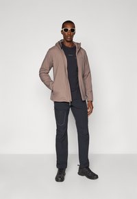 Kevyt, taupe väri zip-up takki hupulla tumman t-paidan päällä, yhdistettynä tummiin, taskullisiin housuihin ja mustiin kenkiin. Valkoiset aurinkolasit.