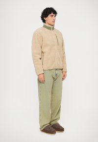 Beige fleecejack met een hoge kraag en groene accenten, gecombineerd met lichtgroene gestreepte broek en donkerbruine instap schoenen met een patroon.