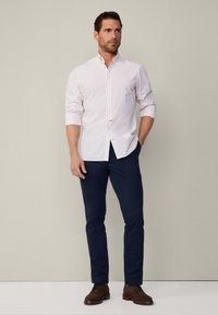 Uomo che indossa una camicia bianca a quadretti rosa con le maniche arrotolate, pantaloni blu navy e scarpe in suede marroni, in piedi contro un muro semplice.