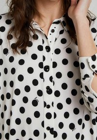 Camicia button-up bianca e nera con grandi pois neri, con colletto e due parole ricamate visibili vicino ai bottoni.