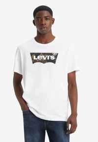 Weißes Baumwoll-T-Shirt mit einem Tarnmuster-Logo von Levi's auf der Vorderseite. Kurze Ärmel und Rundhals-Design. Kombiniert mit dunkelblauen Denim-Jeans.