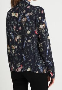Motivo floreale in fiore su una camicia di tessuto blu scuro, con maniche lunghe, colletto alto e polsini con bottoni. Texture liscia.