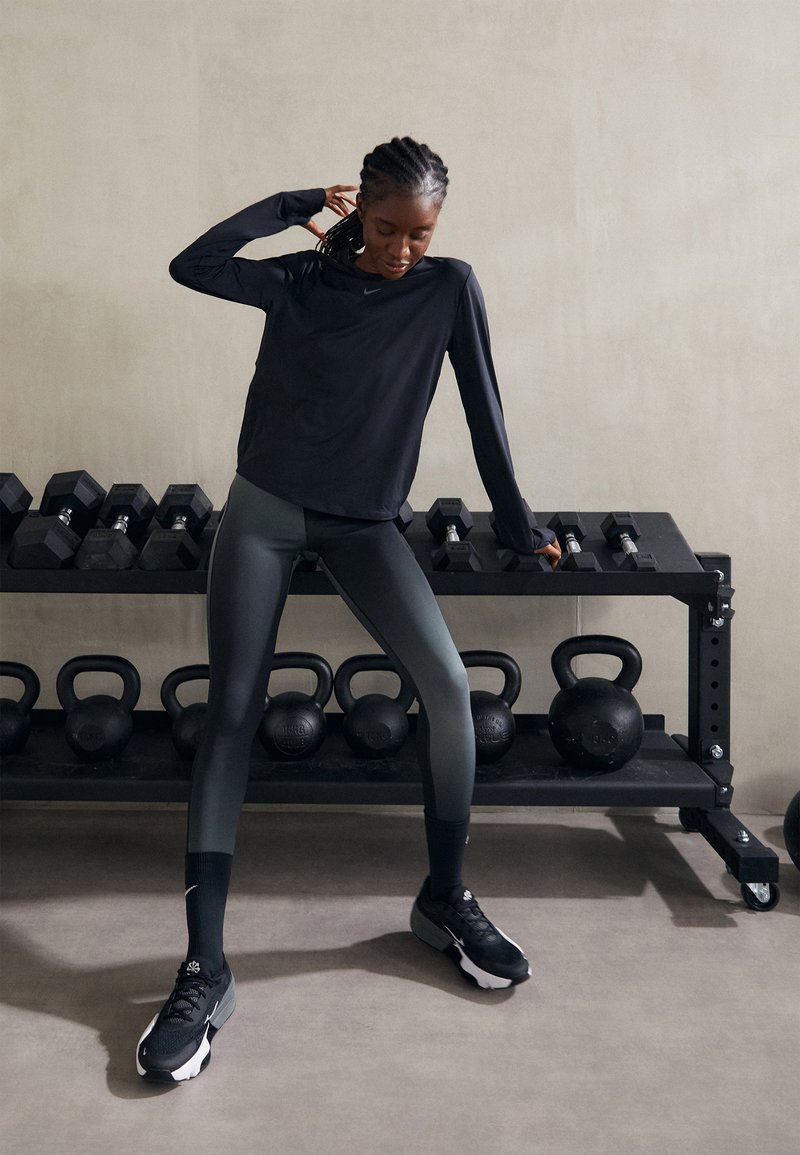 Camisa atlética negra de manga larga, leggings ajustados gris oscuro y zapatillas negras. En el fondo, hay equipos de gimnasio que incluyen pesas y kettlebells.