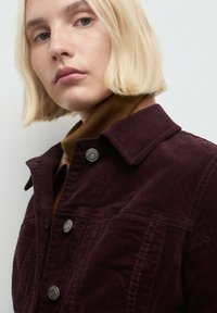 Chaqueta de pana burdeos con cuello, que cuenta con detalles de botones plateados y una tela texturizada. Debajo lleva una sudadera de cuello alto marrón.