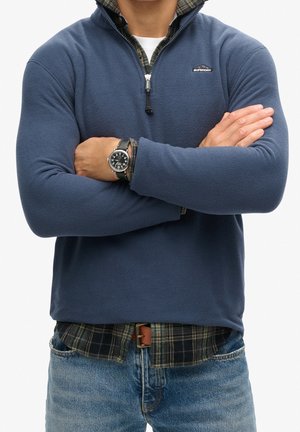 Personne portant une polaire bleue Superdry, les bras croisés, une chemise à carreaux en dessous, un jean bleu et une montre noire au poignet.