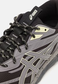 Athletic shoe in black mesh and grey rubber, featuring reflective accents, textured overlays, and patterned laces. Rounded toe design.
