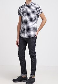 Camisa de cuadros en blanco y negro de manga corta, combinada con jeans oscuros ajustados y zapatos negros con cordones. Tejido suave con un diseño casual.