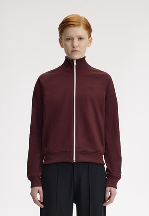 Giovane con capelli rossi che indossa una giacca sportiva color bordeaux con cerniera e pantaloni neri, in piedi davanti a uno sfondo bianco uniforme.