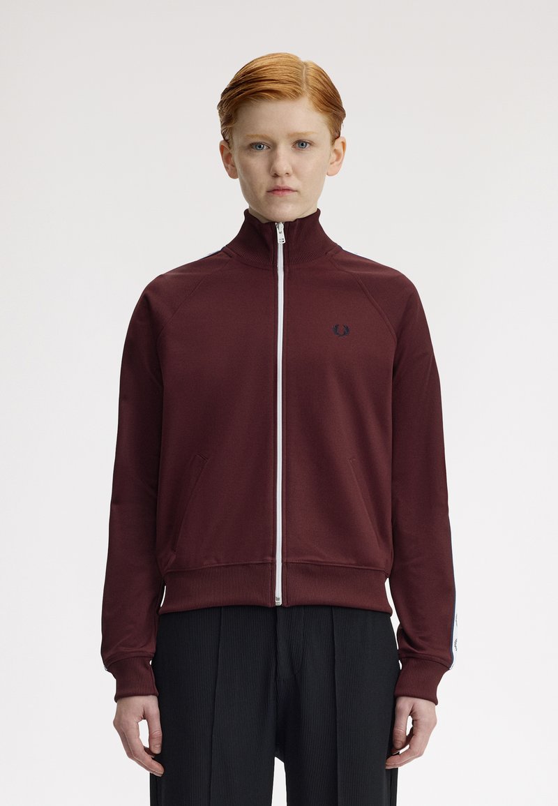 Ung person med rødt hår iført en lynlåst bordeaux træningsjakke og sorte bukser, stående mod en ensfarvet hvid baggrund.