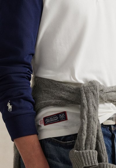 Chemise à manches longues marine avec emblème, polo blanc en dessous et un pull gris texturé noué à la taille, avec une étiquette.