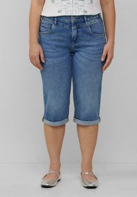 Un pantalon capri en denim bleu clair avec des ourlets roulés et des poches avant. Porté avec un T-shirt graphique blanc et des chaussures plates argentées.