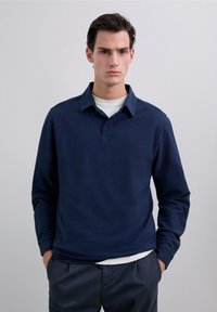 Camisa polo de manga larga azul marino hecha de tejido suave, con un cuello clásico, tapeta de botones y un diseño minimalista. Combinada con una camiseta blanca debajo.