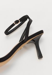 Sandale noire à talons, présentant un design élégant de brides en cuir, une bride de cheville avec une boucle dorée et un talon fin et haut pour apporter de la hauteur.