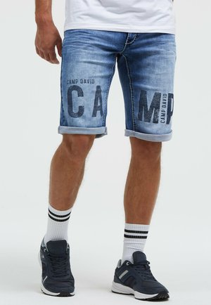 Man wearing rolled-up blue denim shorts with "CAMP DAVID" text, white socks with black stripes, and black athletic shoes.