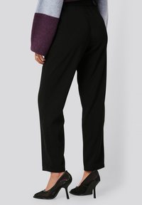 Černé kalhoty na míru s hladkou texturou, spárované s černými síťovanými lodičkami na vysokém podpatku s špičatou špičkou a stiletto podpatkem.