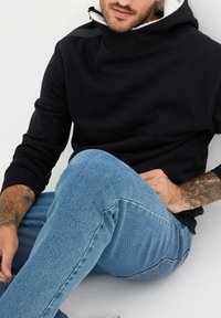 Felpa nera con cappuccio e collo alto, realizzata in materiale morbido. Abbinata a jeans in denim blu, caratterizzati da una texture sbiadita e un design aderente.